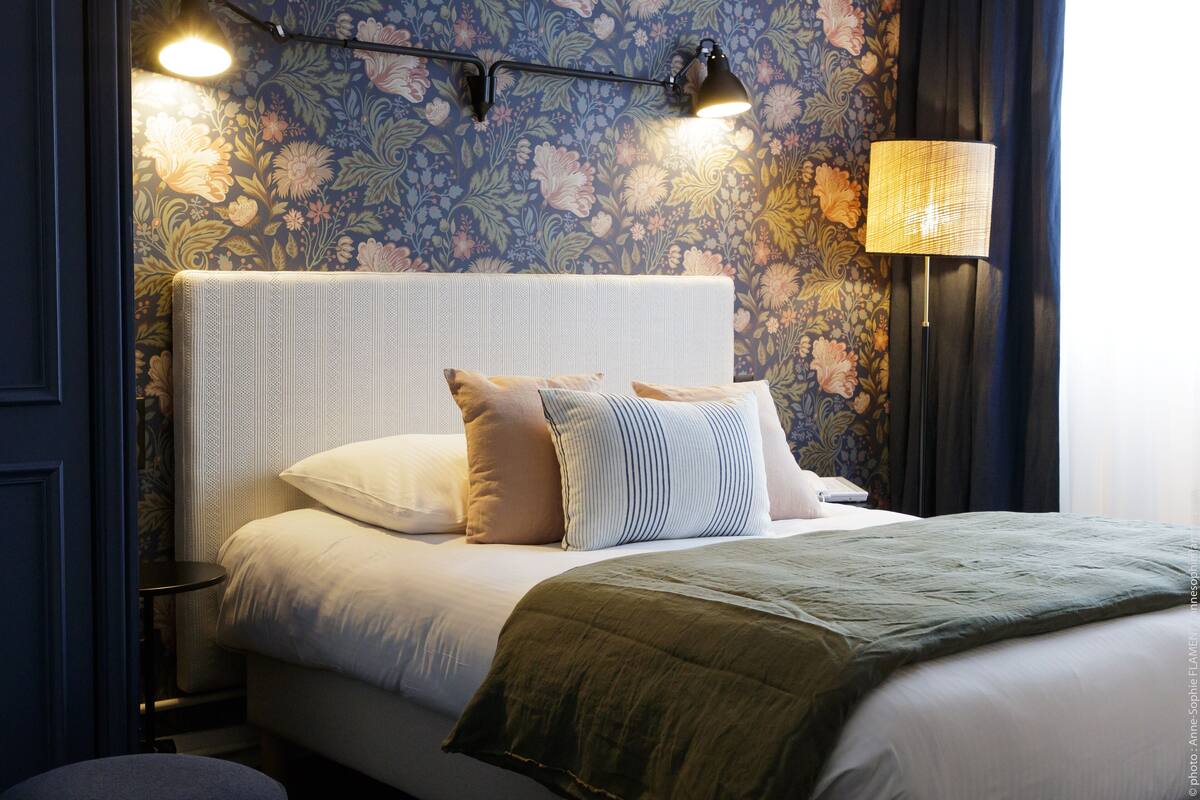 Ein ordentlich bezogenes Bett mit grüner Tagesdecke, ein Kopfteil mit Blumenmuster und eine Wandlampe.