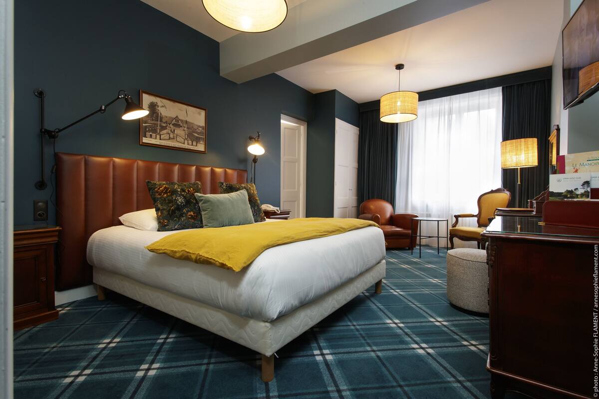 Ein Hotelzimmer mit einem großen Bett, einem Kopfteil, einem Nachttisch, einem Sessel, einem Schreibtisch und einem Fernseher.