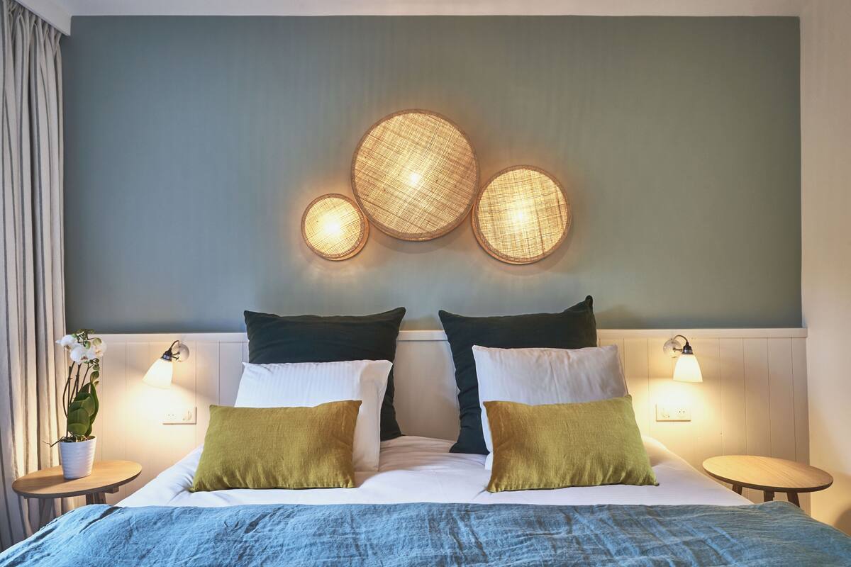 Ein ordentlich bezogenes Bett mit weißen und grünen Kissen, einer blauen Decke und einer Wandleuchte darüber.