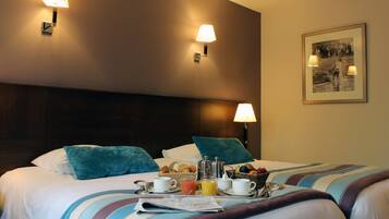 A hotel room with a neatly made bed, breakfast tray with croissants, fruit, and beverages, and two wall-mounted lamps.