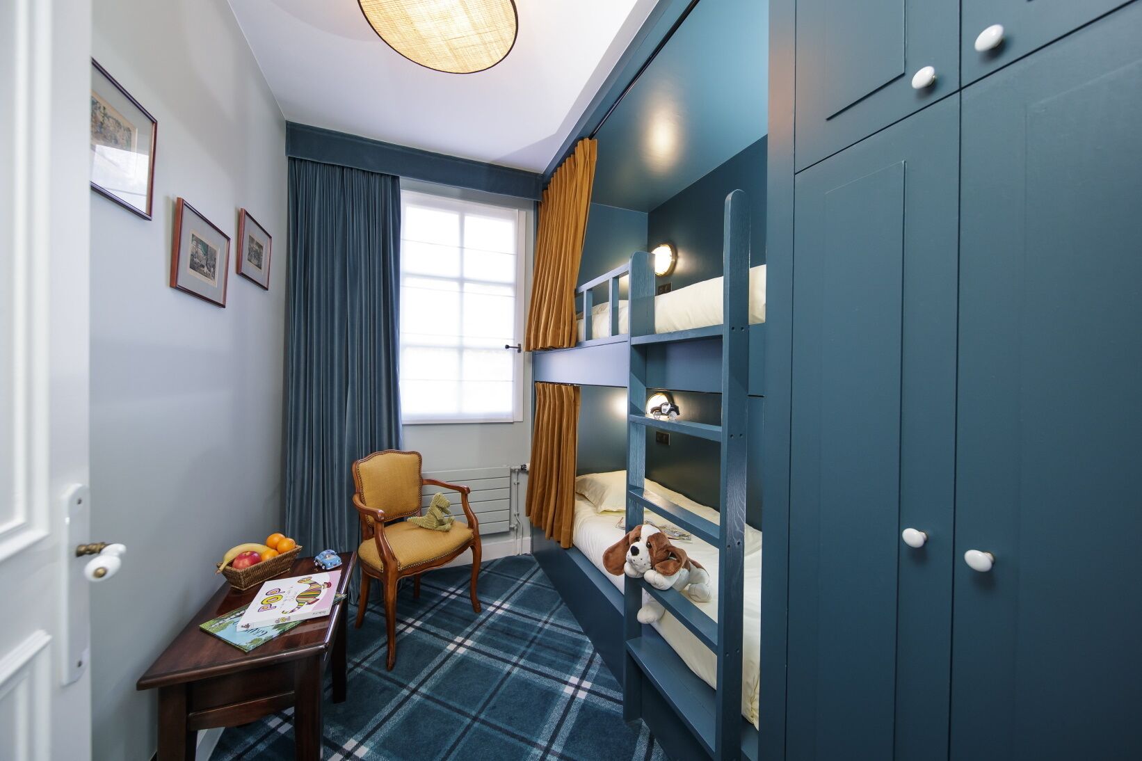 Ein Zimmer mit einem Etagenbett, einem Sessel, einem kleinen Tisch mit Büchern und Obst sowie einem Fenster mit Vorhängen.