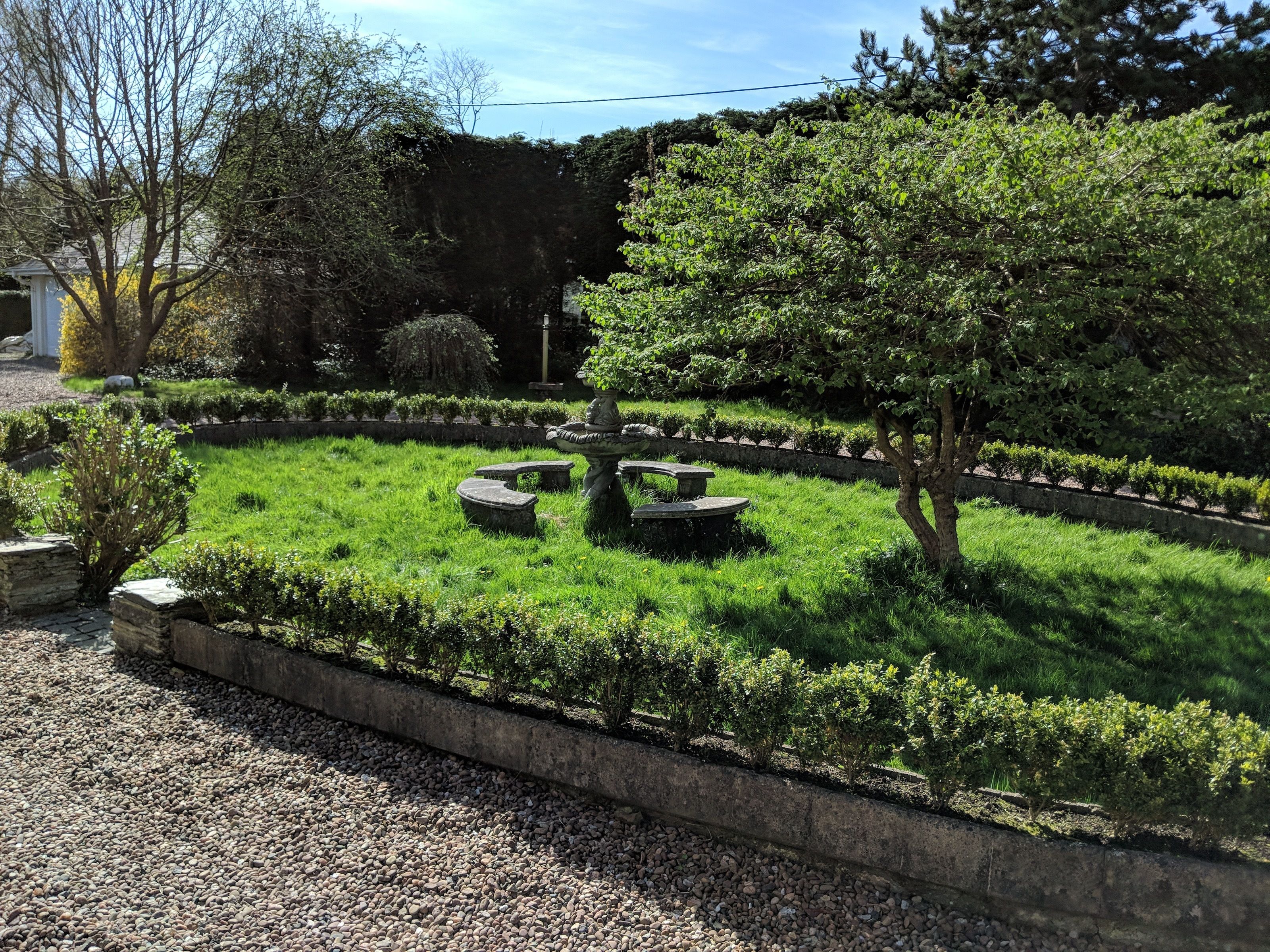 A well-maintained garden with a circular stone seating area, surrounded by neatly trimmed hedges and a variety of trees.