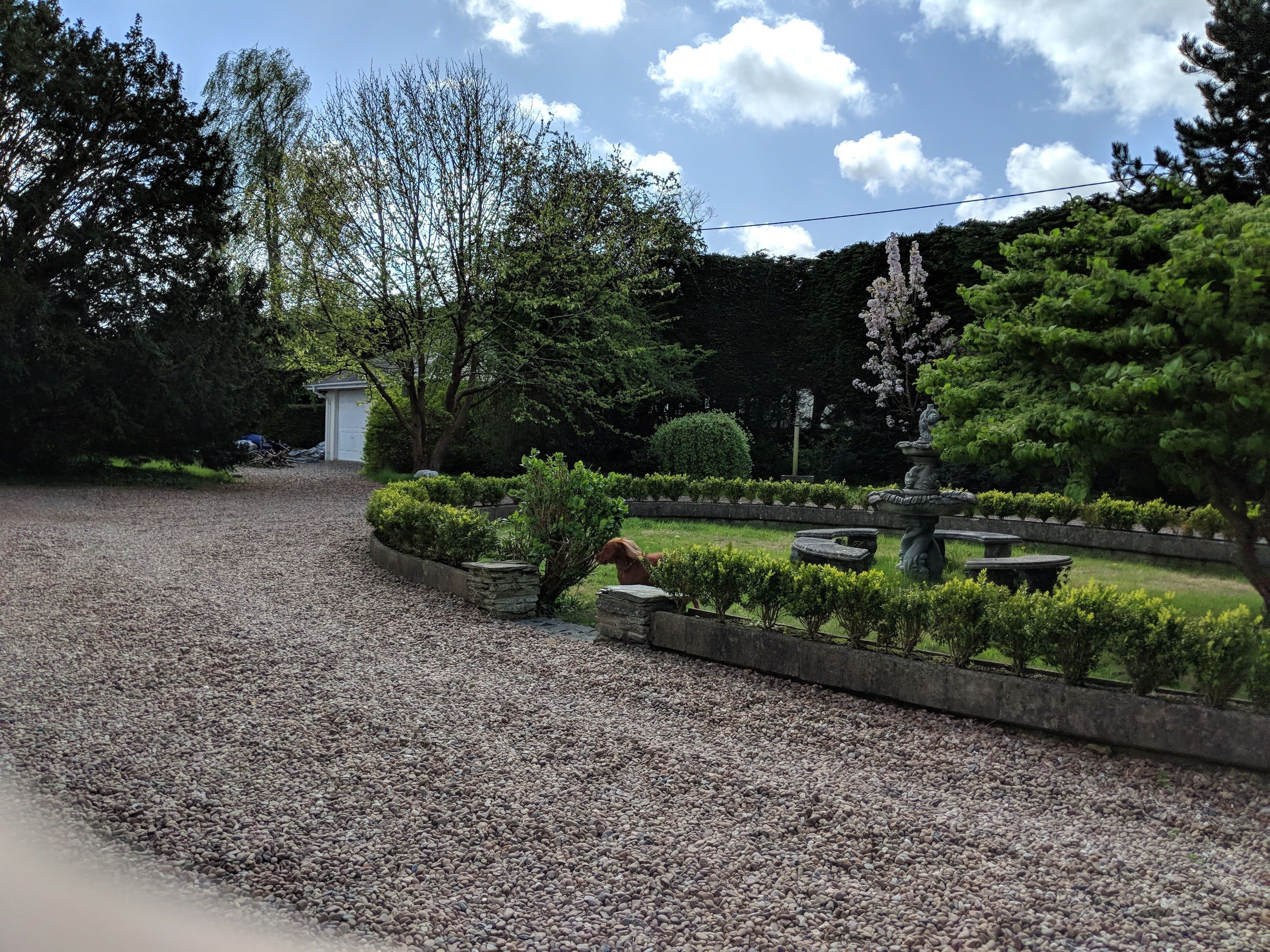 Un camino de grava conduce a un jardín bien cuidado con una estatua, setos cuidadosamente recortados y un pequeño cobertizo al fondo.