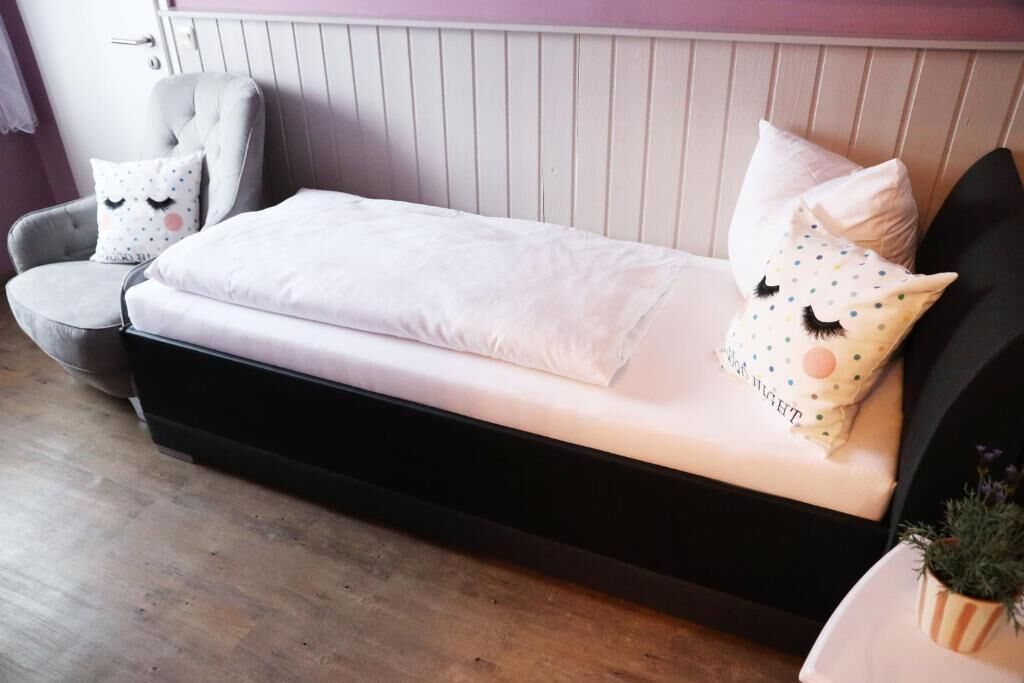 Una cama con colchón blanco y estructura negra, un sillón gris con almohada blanca, y una pequeña planta en maceta a la derecha.