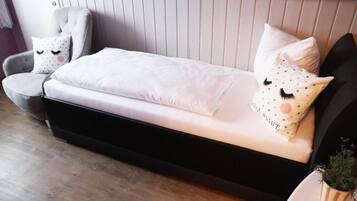 Una cama con colchón blanco y estructura negra, un sillón gris con almohada blanca, y una pequeña planta en maceta a la derecha.