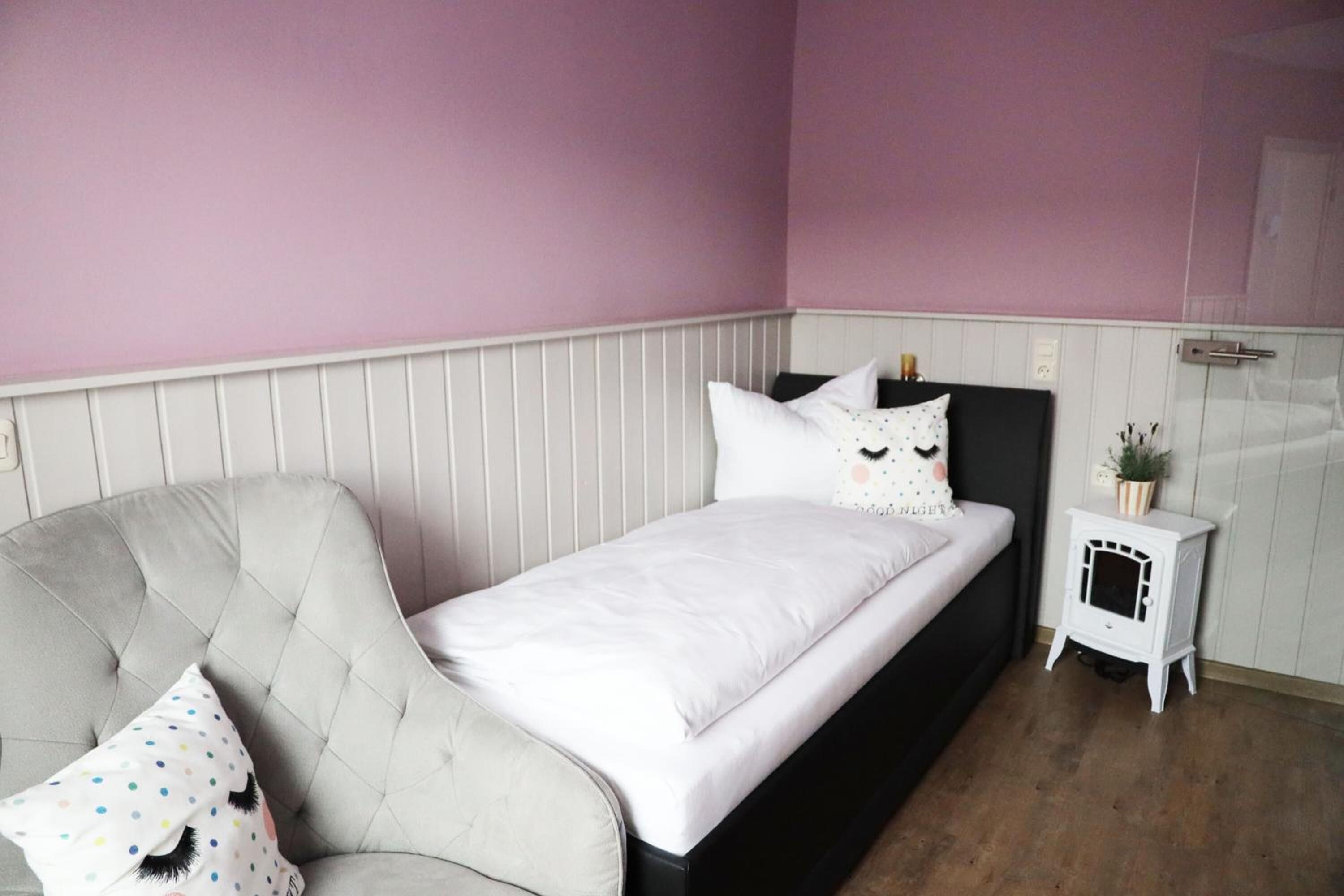 A bedroom with a bed, a chair, a small white heater, and a wall-mounted shelf.