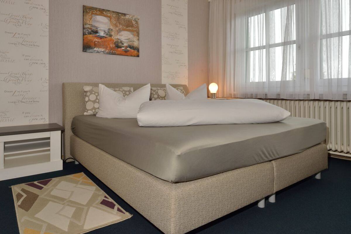 Uma cama bem arrumada com colchão e almofadas bege, uma mesinha de cabeceira com luminária e uma janela com cortinas leves.