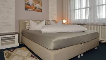 A neatly made bed with a beige mattress and pillows, a bedside table with a lamp, and a window with sheer curtains.