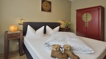 Una cama bien hecha con una colcha blanca y almohadas, un par de botas sobre la cama, un reloj de pared y un adorno decorativo en la pared.