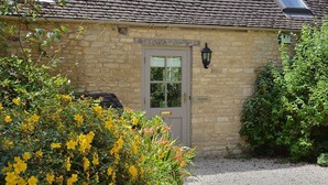 A stone building with a door labeled "Shipton," surrounded by greenery and a gravel path. - Shipton Cottage (Chipping Norton)
