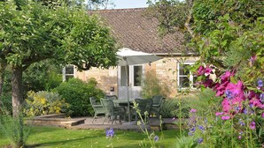 A stone building with a tiled roof, a garden with a table and chairs, and flowering plants. - Shipton Cottage (Chipping Norton)