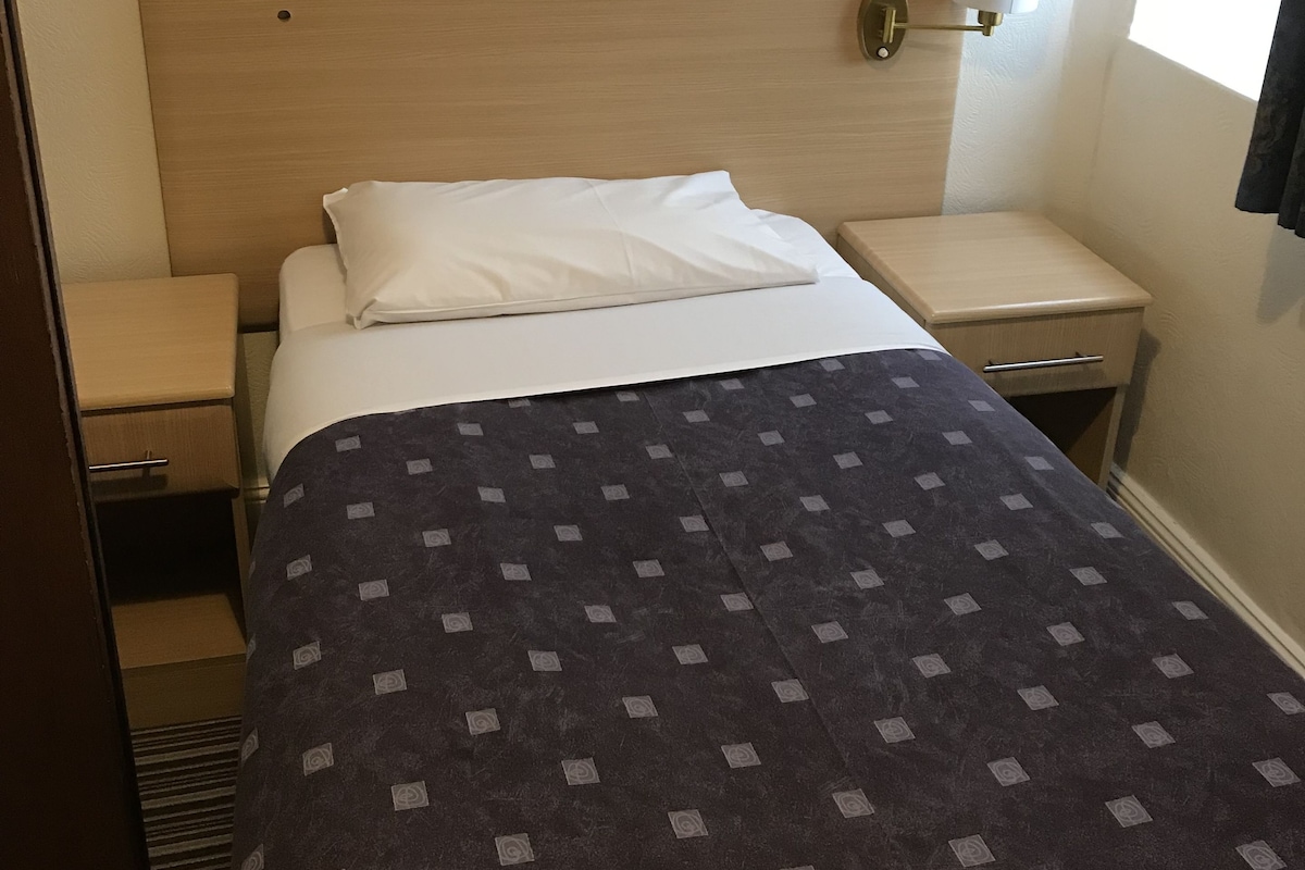 Una habitación de hotel con una cama, una mesita de noche, una lámpara y una ventana.