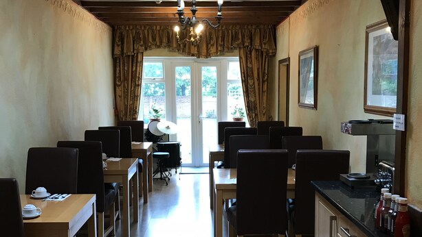 A dining area with wooden tables and chairs, a kitchen area with a black countertop, and a view through a doorway to an outdoor space. - Warwick Lodge (Surbiton)