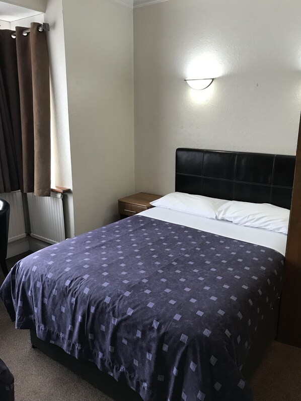 A hotel room with a bed, a nightstand, a lamp, and a window with curtains. - Warwick Lodge (Surbiton)