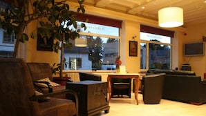 Un salon chaleureux avec un canapé, un fauteuil et une petite table. De grandes fenêtres sont ornées de rideaux, et on y trouve une plante en pot et une lampe.