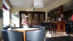 Un espace bar avec un comptoir et des tabourets, un bureau d’accueil agrémenté d’un vase de fleurs et un coin salon avec des fauteuils en cuir.