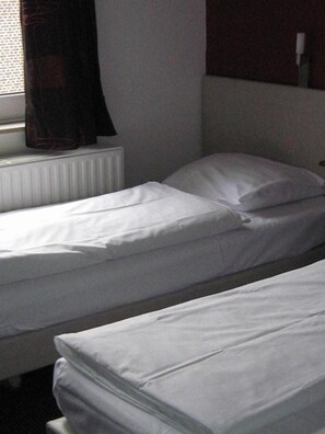 Una cama individual con ropa de cama blanca en una habitación con ventana y radiador.