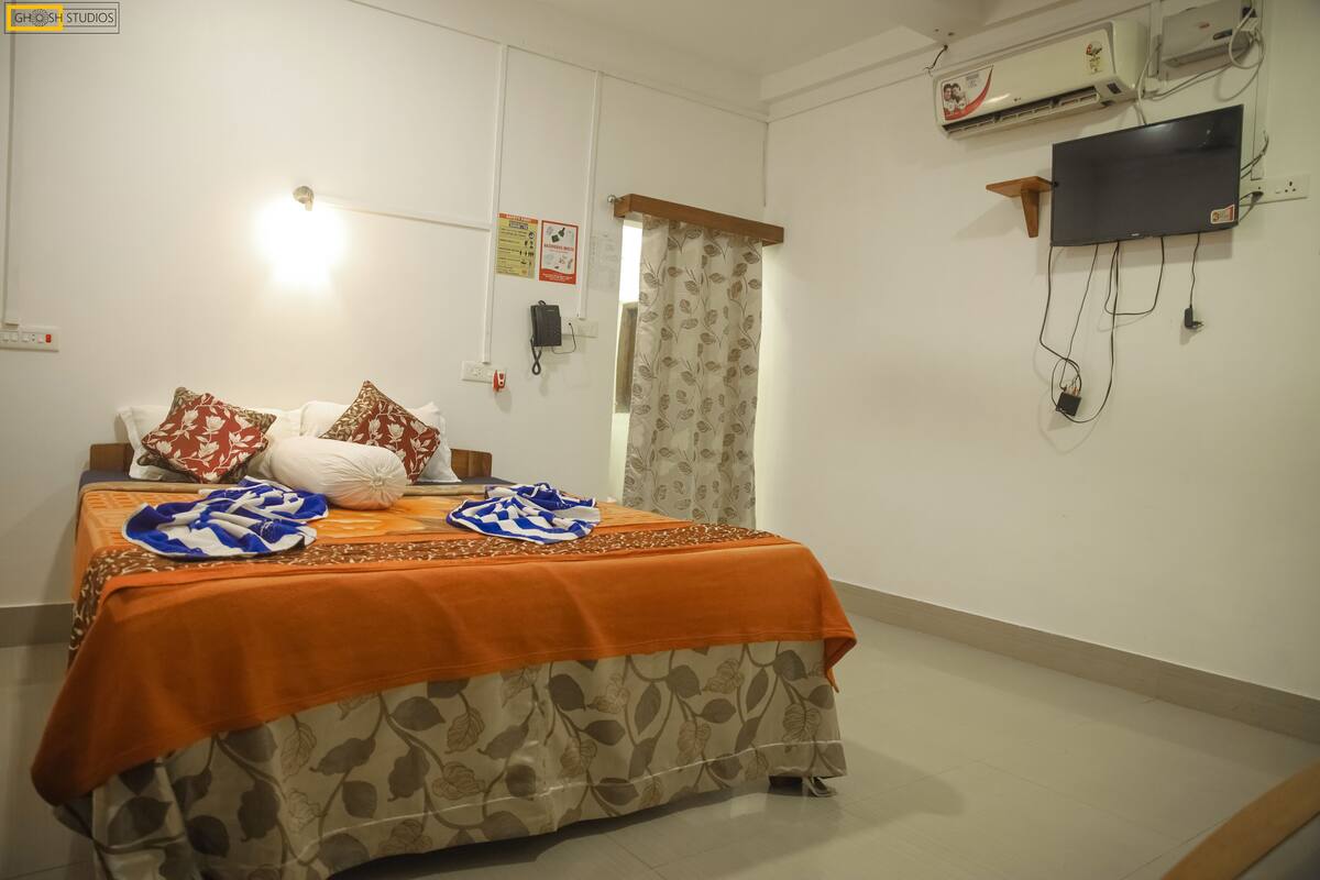 A hotel room with a bed, a flat-screen TV, an air conditioning unit, and a wall-mounted phone.