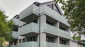 Un immeuble moderne à plusieurs étages, doté de balcons, de grandes fenêtres et d’un espace jardin agrémenté de diverses plantes.