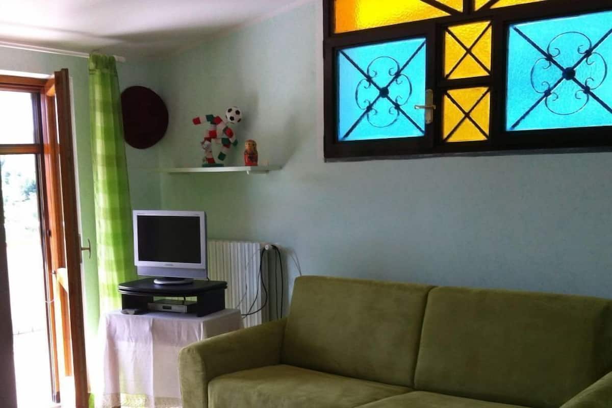 Een woonkamer met een groene bank, een televisie op een standaard, een klein tafeltje en een raam met decoratieve glaspanelen.