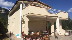 Una casa con porche techado, mobiliario exterior y montañas al fondo.