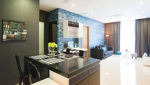 A modern kitchen with a central island, a dining area with a black table and chairs, a living area with a sofa and a TV, and a ceiling fan.