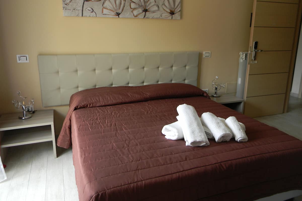 A neatly made bed with a brown bedspread and white towels, a tufted headboard, a bedside table with a lamp, and a wall with a floral painting.