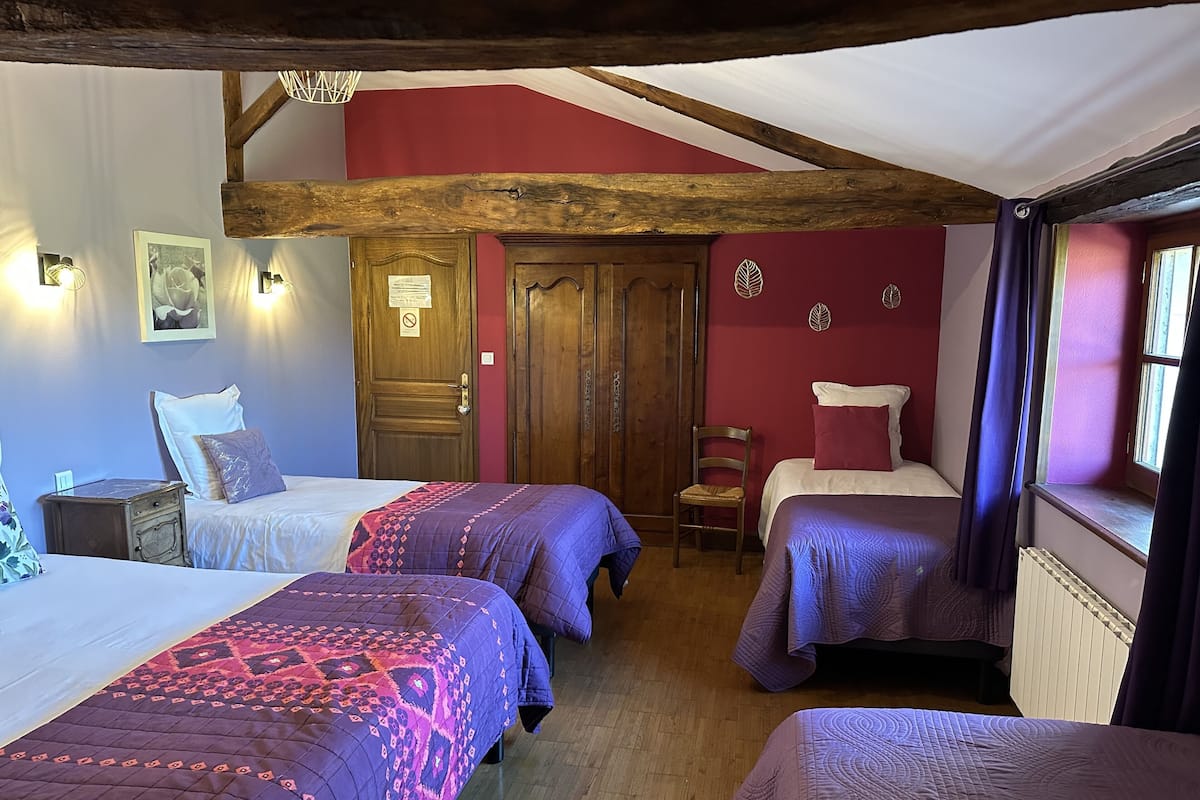 A room with two beds, wooden beams, a red wall, and a window with curtains.
