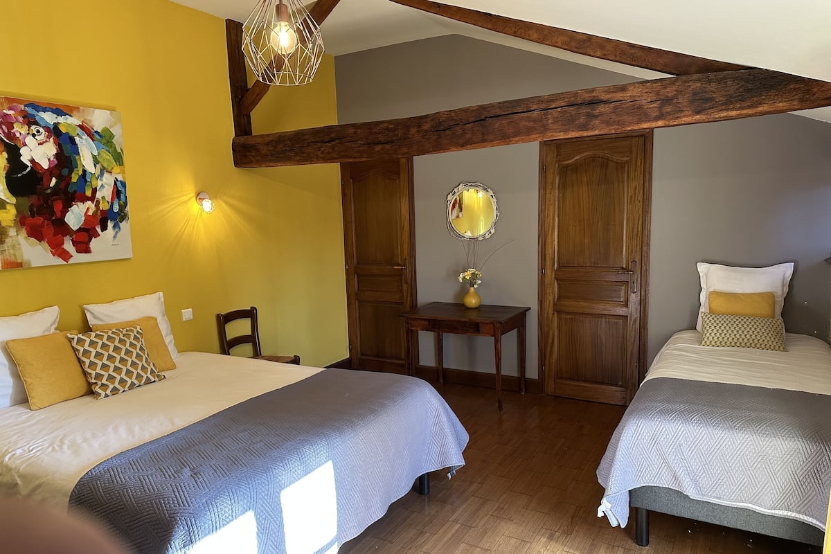 A bedroom with two beds, wooden beams, a painting, a mirror, and a small table with a vase.