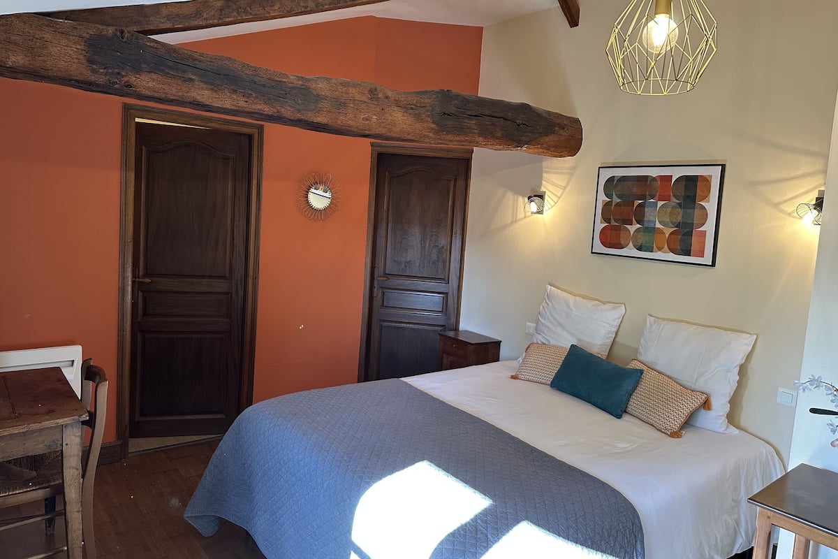 A bedroom with a bed, wooden beams, a wall clock, a geometric light fixture, and a framed picture.