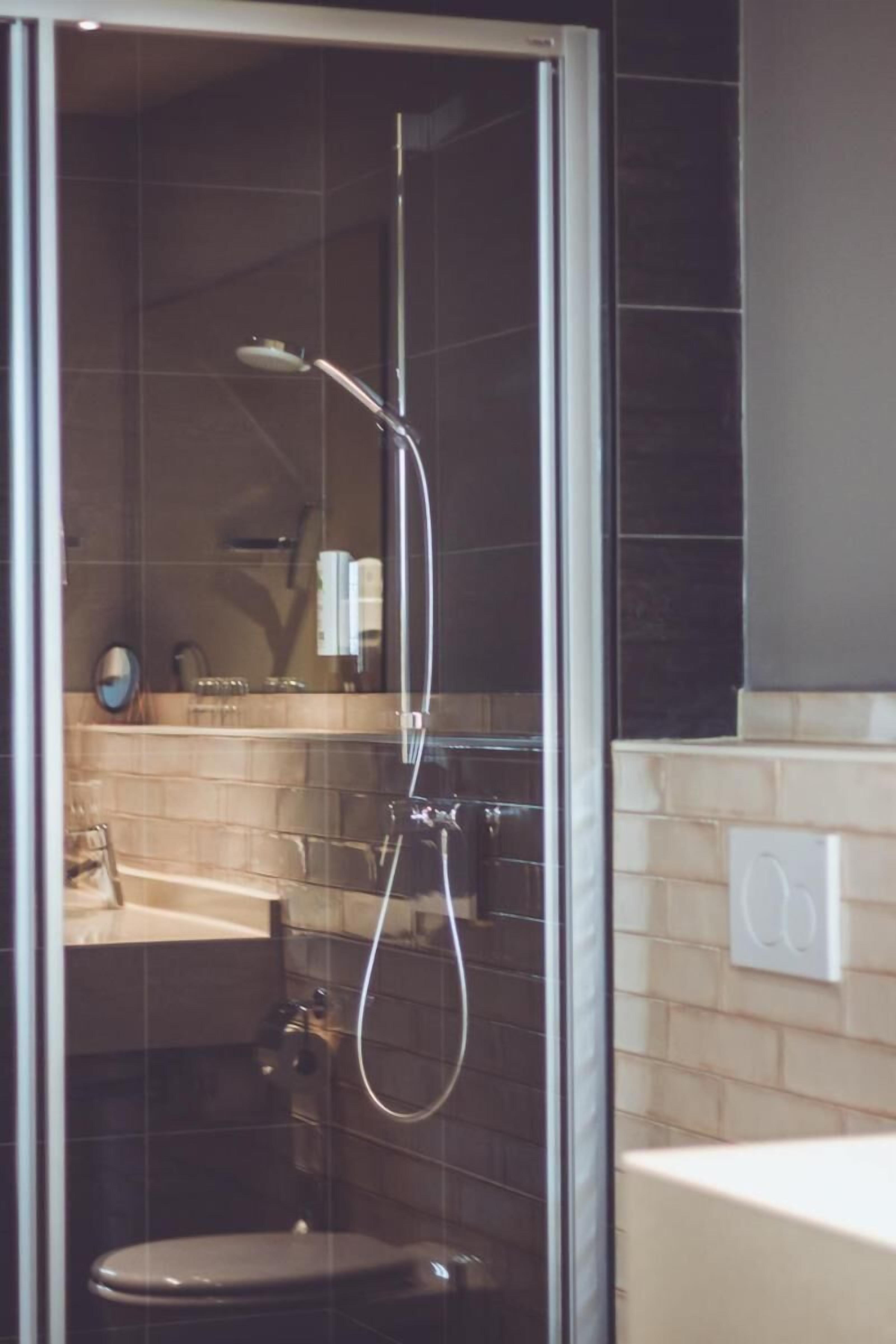 A glass-enclosed shower area with a handheld showerhead, a toilet, and a sink visible through the glass.
