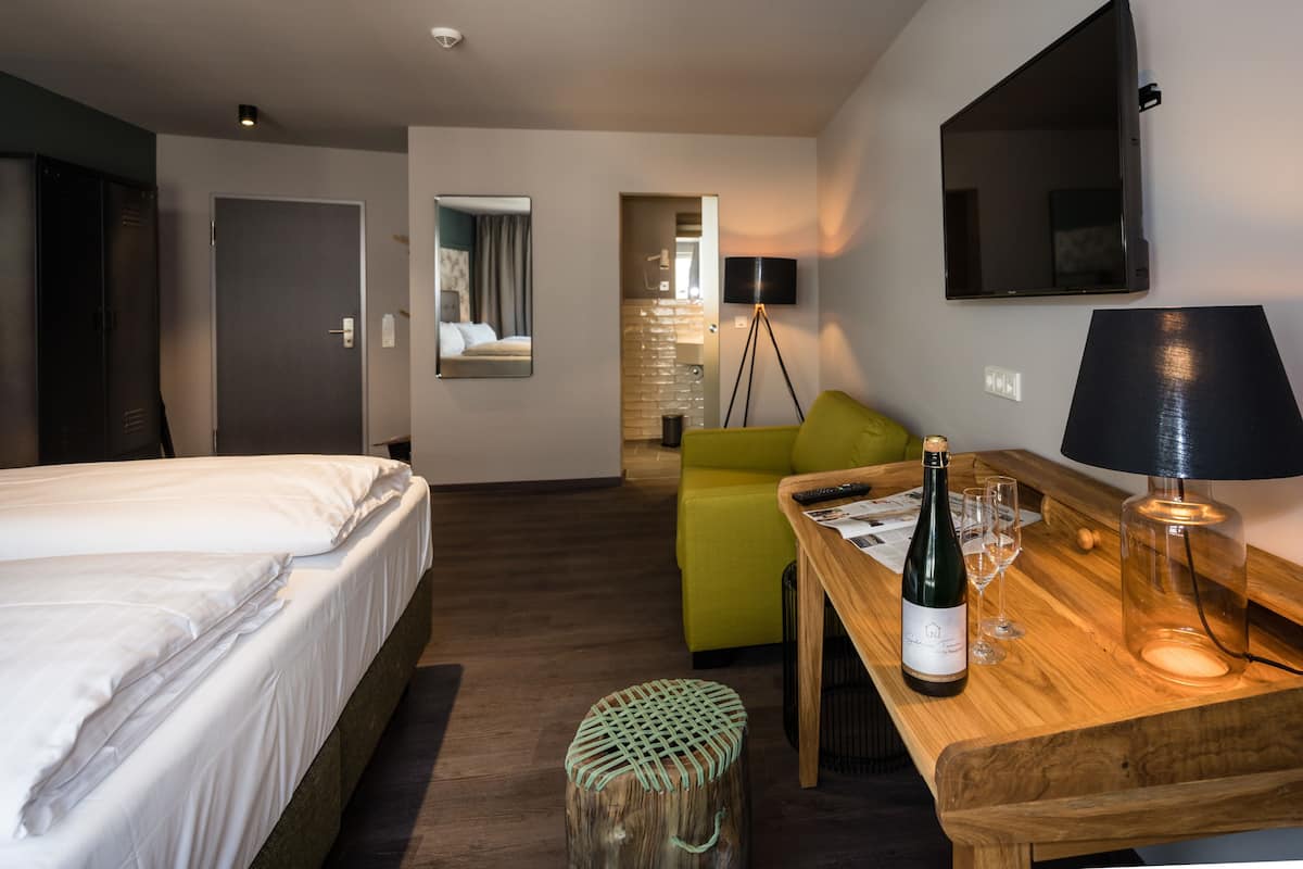 Une chambre d’hôtel avec un lit, une table en bois sur laquelle se trouvent une bouteille et des verres, un fauteuil vert, un miroir et un téléviseur fixé au mur.
