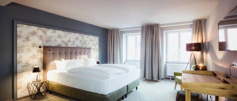 Habitación de hotel con una cama grande, un escritorio y una silla. Tiene una ventana con cortinas, una lámpara y un televisor montado en la pared.