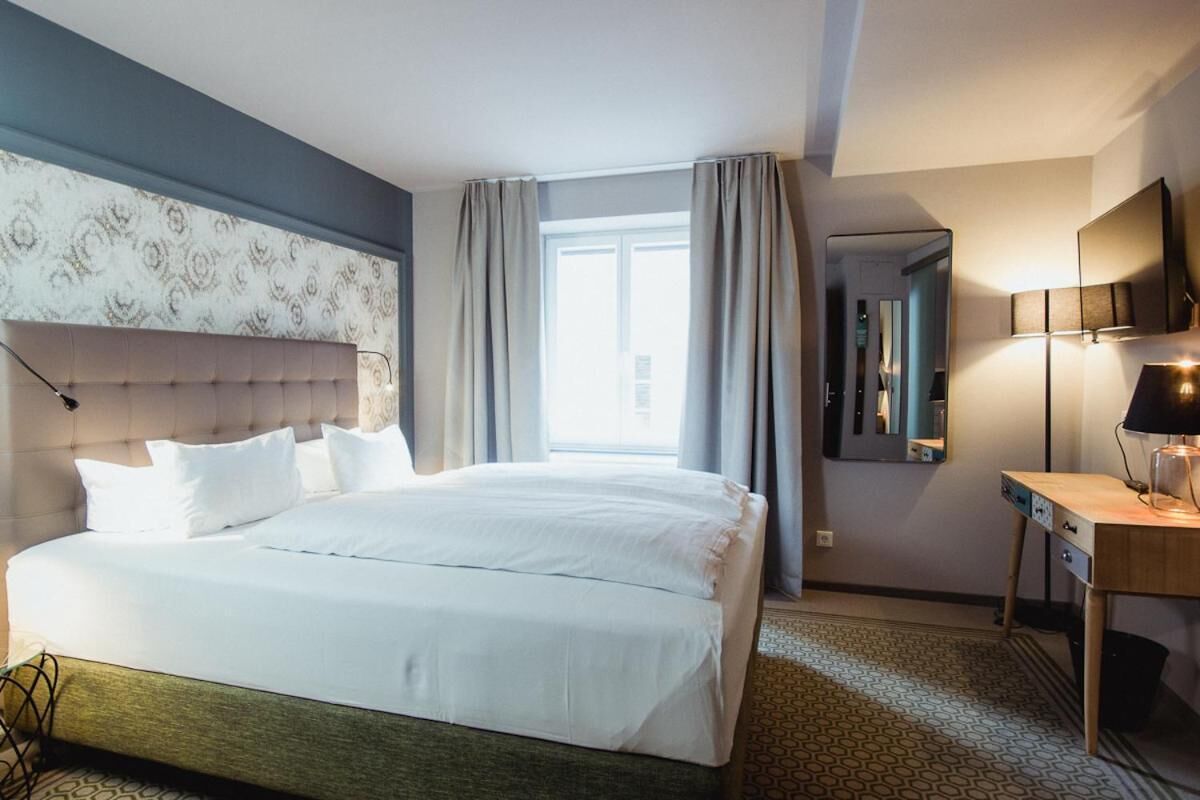 A hotel room with a large bed, a desk with a lamp, a TV, and a window with curtains.