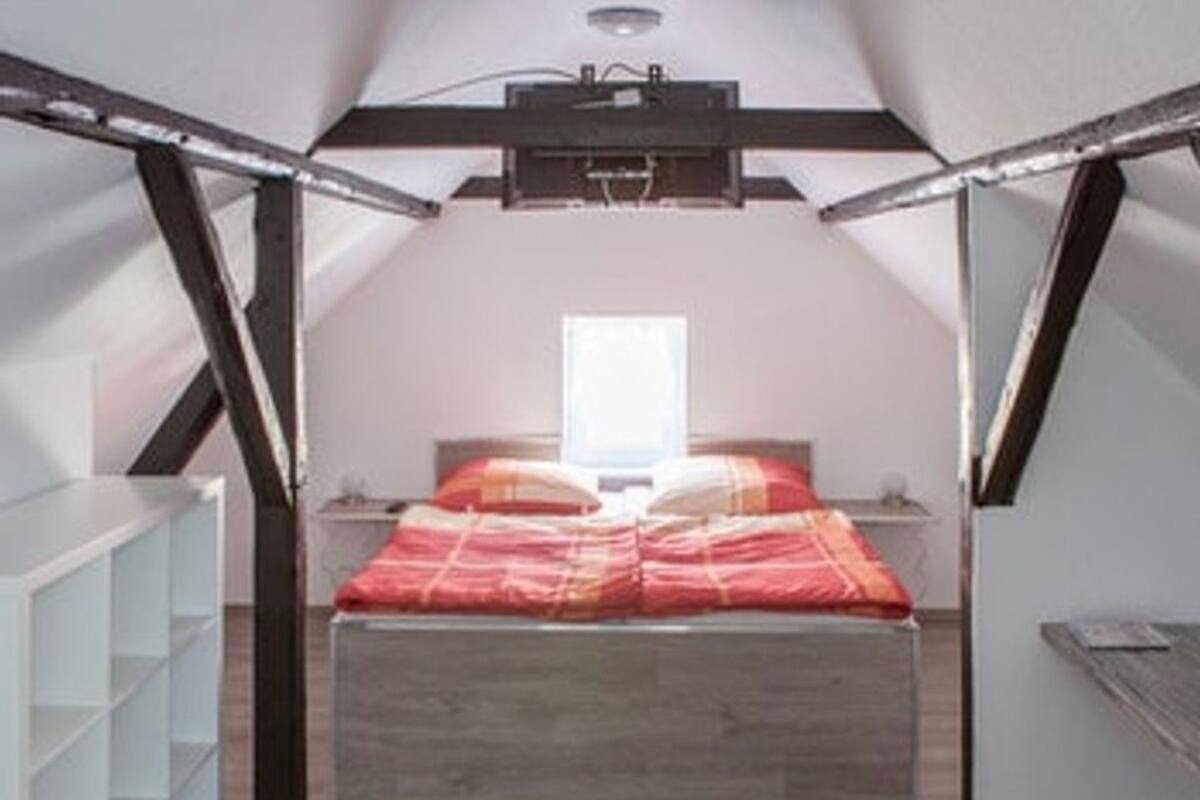 A bedroom with a sloped ceiling, a bed with a red and white bedspread, a black rug, and a white shelving unit.