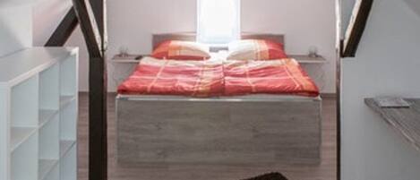 Un dormitorio con techo inclinado, una cama con colcha roja y blanca, un tapete negro y un estante blanco.