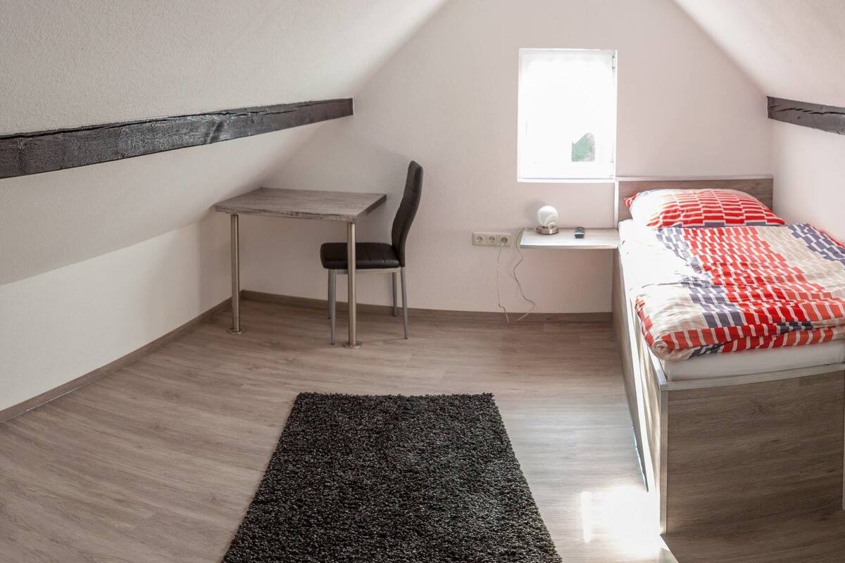 A small, well-lit room with a sloped ceiling, a desk with a chair, a bed with a patterned blanket, and a white shelving unit.