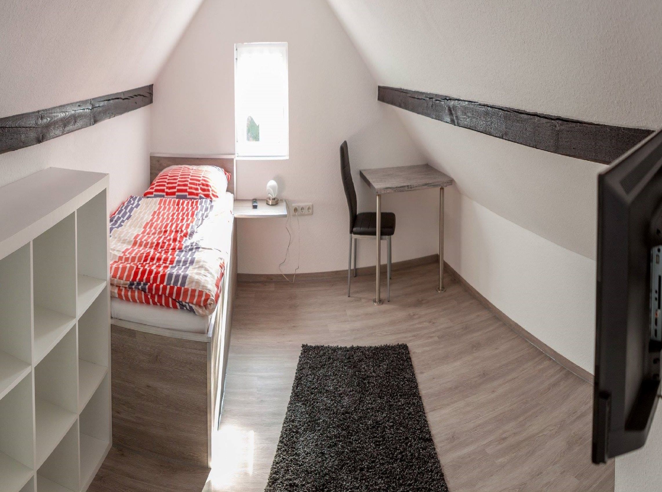 A small, well-lit room with a bed, a desk, a chair, and a TV.