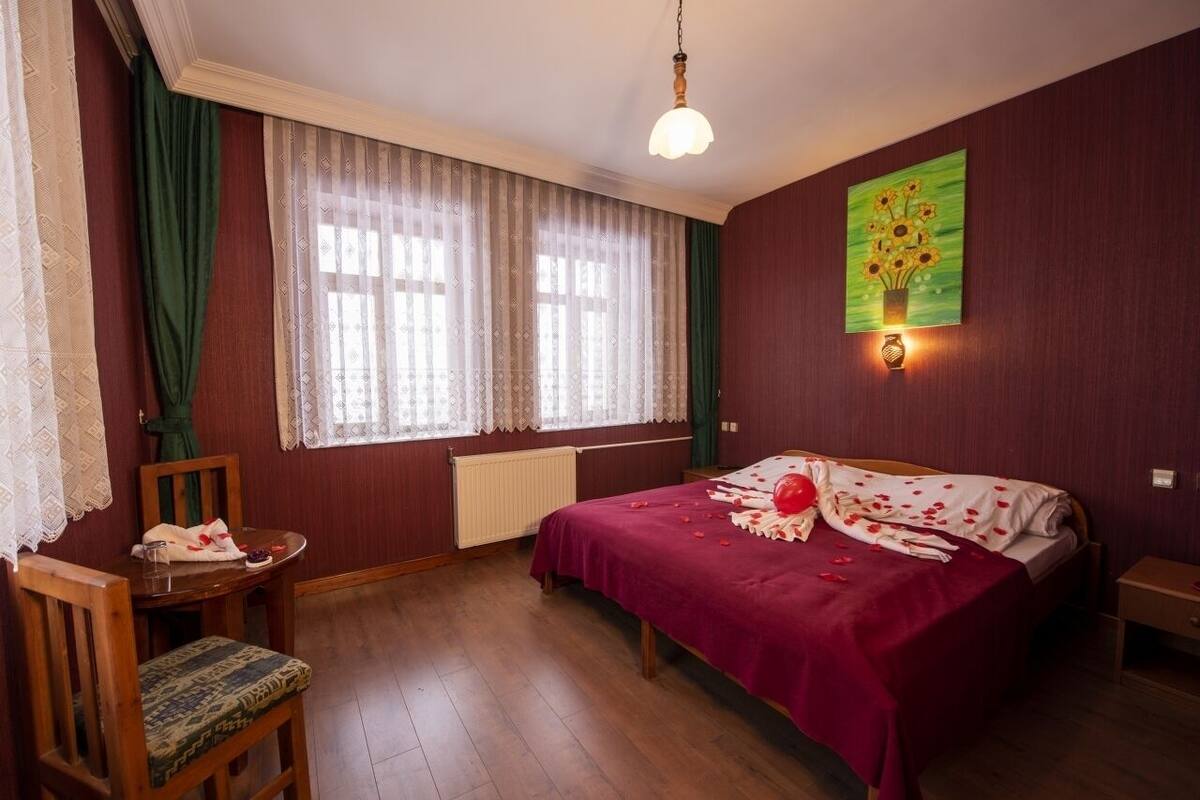 A bedroom with a bed, a chair, a table, a window with curtains, and a wall-mounted light fixture.
