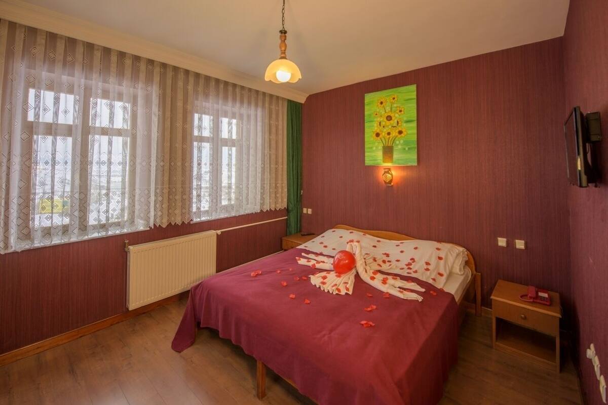 A bedroom with a bed, a window with curtains, a radiator, a bedside table, and a wall-mounted TV.