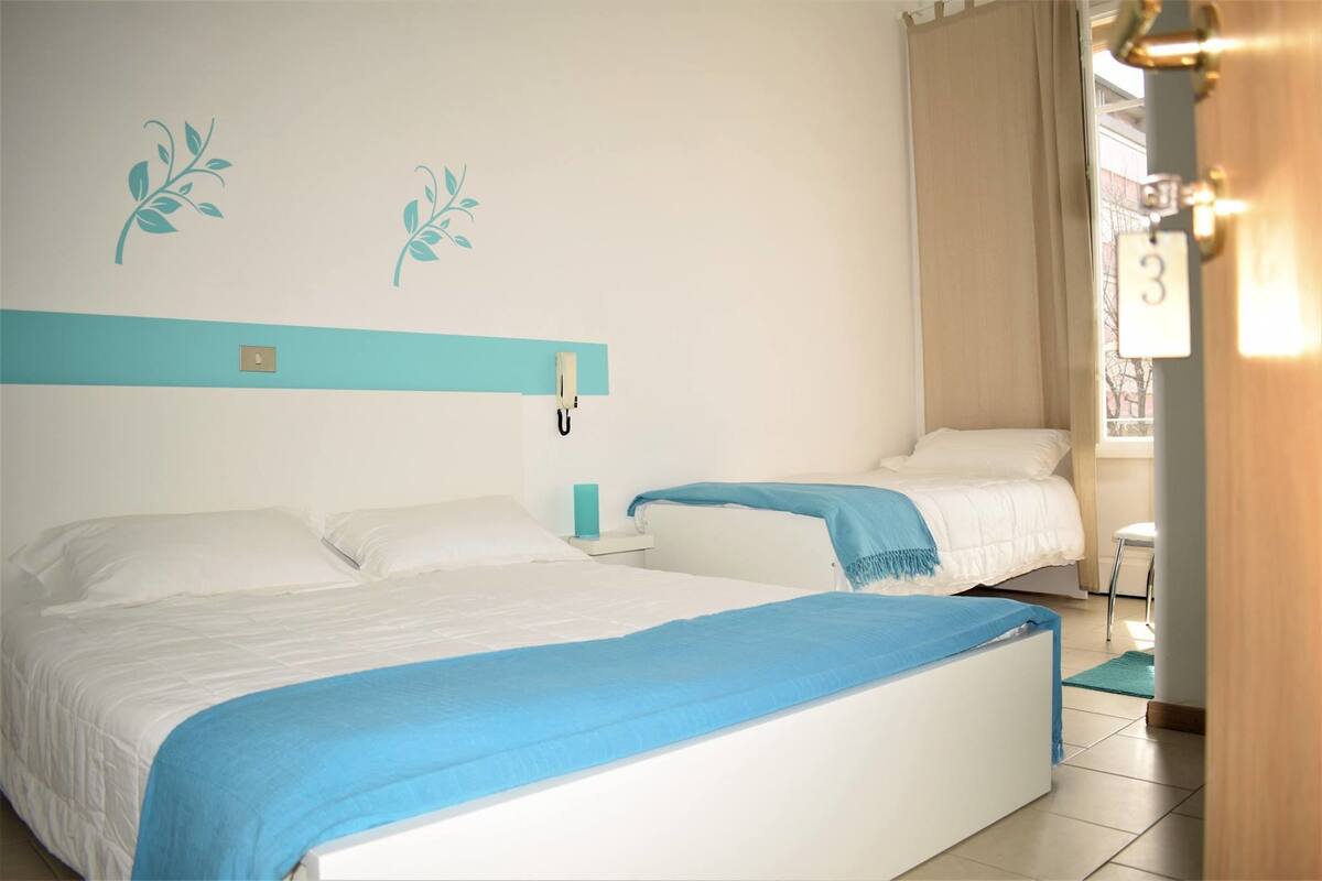 A hotel room with two beds, a nightstand, a lamp, and a view of the outside.