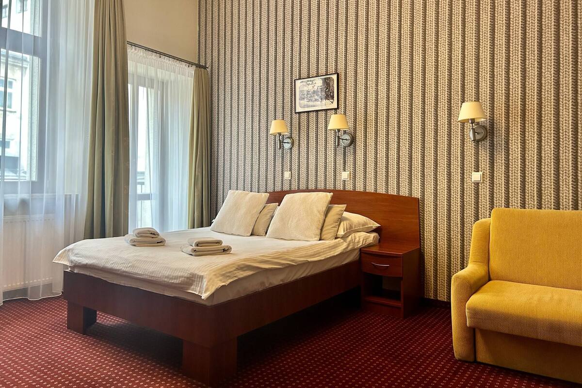 A hotel room with a bed, a chair, a nightstand, a lamp, and a wall with a patterned wallpaper.