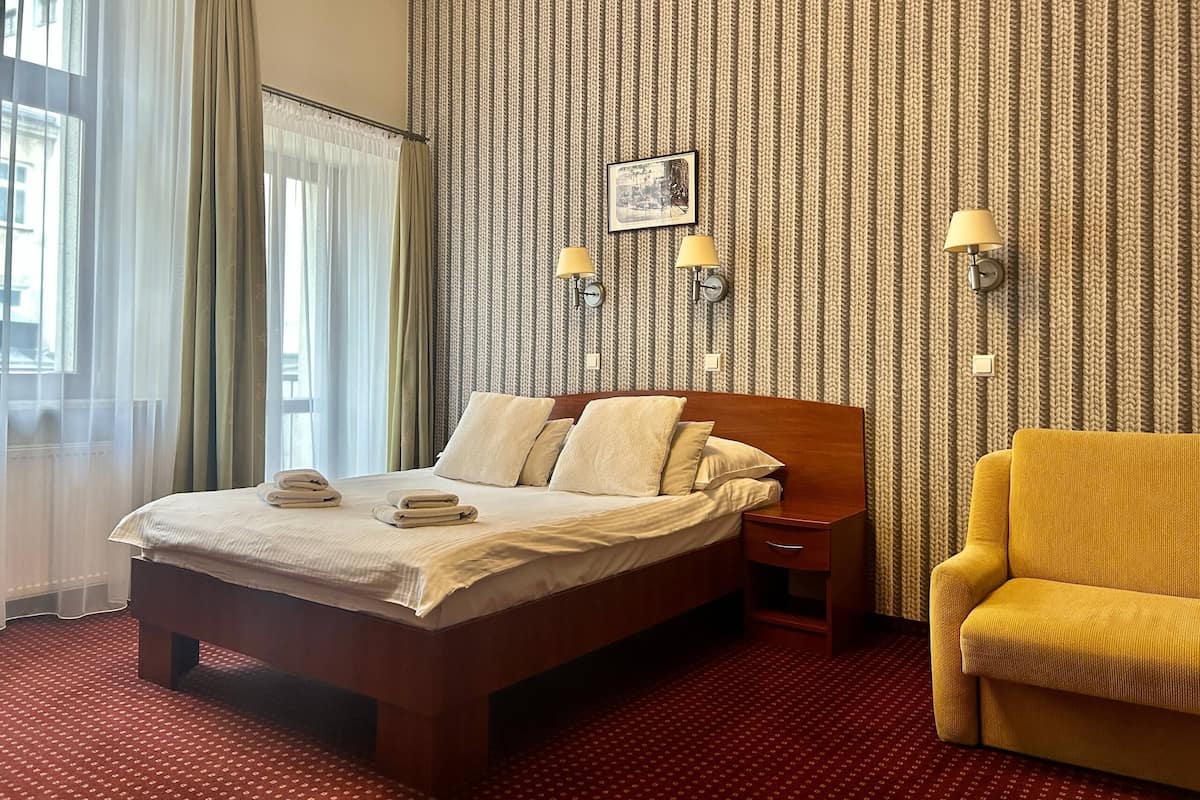 Ett hotellrum med en säng, en stol, ett nattduksbord, en lampa och en vägg med tapet med ett mönster.