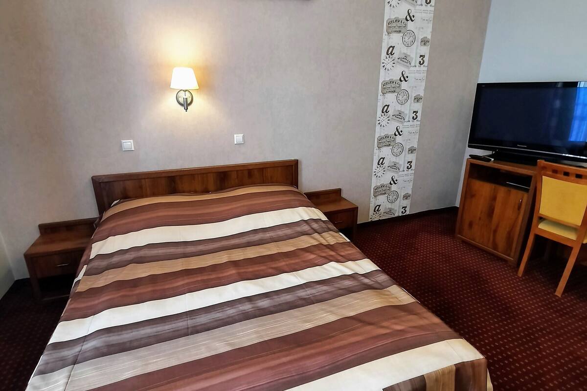 A hotel room with a bed, a TV, a nightstand, a lamp, and a wall with a clock and a floral painting.