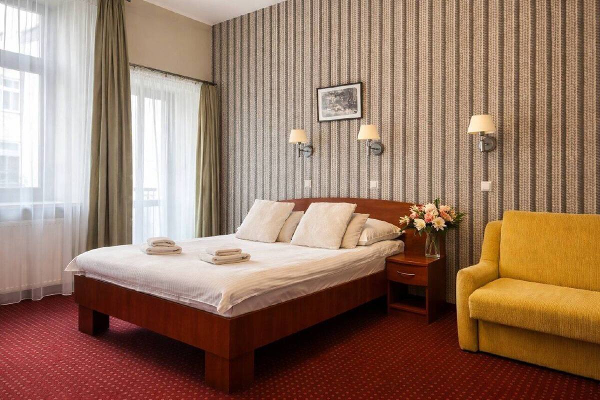 A hotel room with a large bed, a nightstand, a yellow armchair, and a patterned wallpaper.