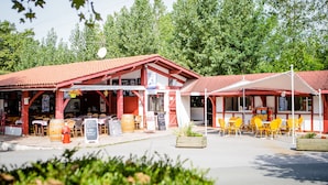 Un área de estar al aire libre de estilo rústico con mesas de madera y sillas amarillas, un bar de aperitivos y un edificio con techo rojo que muestra un letrero que dice "SNACK BAR".