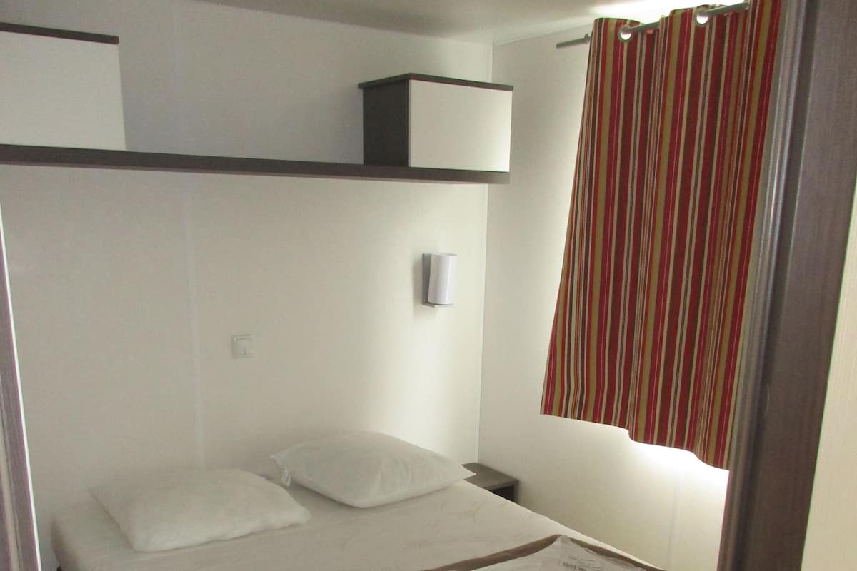 A single bed with a white mattress and pillows, a small bedside table with a lamp, and a striped curtain.