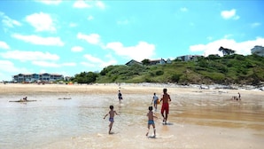 Una playa con gente caminando y jugando en el agua. Se ven casas al fondo, en una colina.
