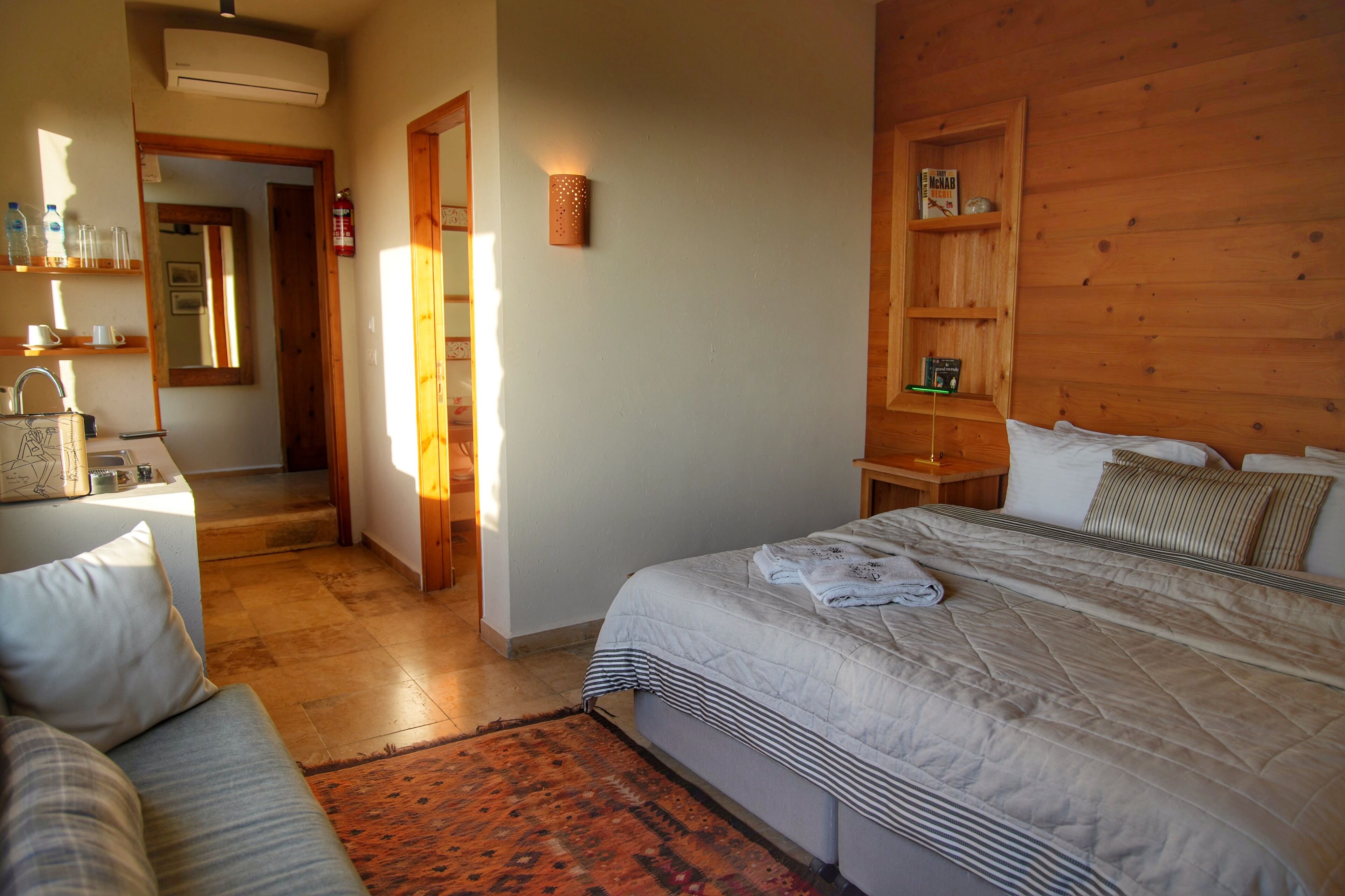 A bedroom with a large bed, a wooden headboard, a bedside table, a nightstand, and a small shelf with books and a lamp.
