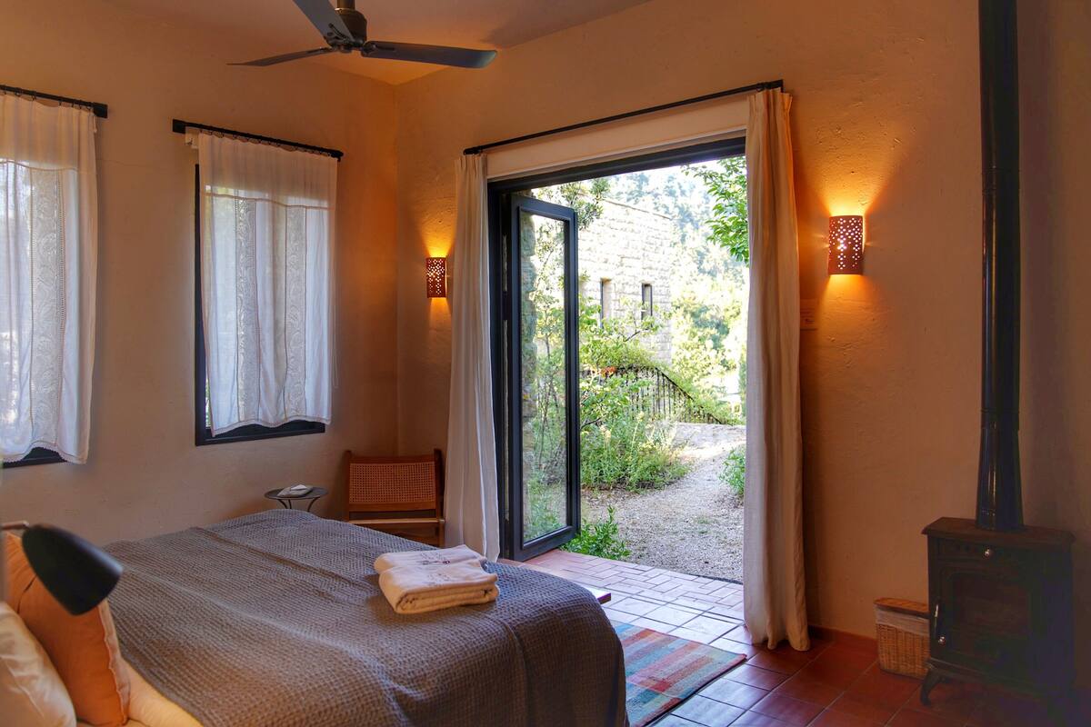Ein Schlafzimmer mit Bett, Deckenventilator, Fenster mit Vorhängen, Nachttisch und Blick ins Freie.